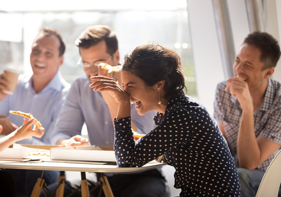 Indian woman laughing at funny joke eating pizza with diverse coworkers in office, friendly work team enjoying positive emotions and lunch together, happy colleagues staff group having fun at break